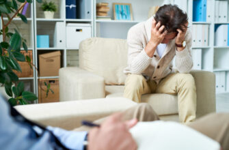 Мужчина с выражением беспокойства сидит на диване, держась за голову. На переднем плане видна рука человека, пишущего заметки.