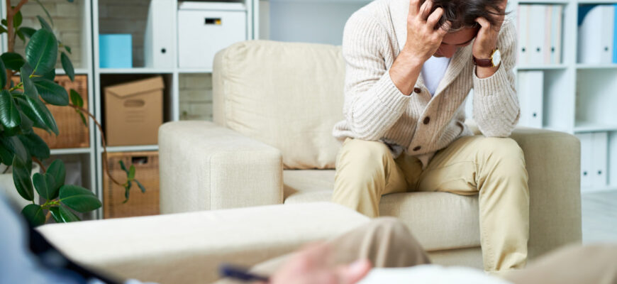 Мужчина с выражением беспокойства сидит на диване, держась за голову. На переднем плане видна рука человека, пишущего заметки.