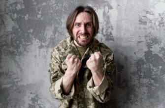 Мужчина в военной форме с длинными волосами выражает гнев, сжатые кулаки подняты к груди. Фон изображает текстурированную серую стену.